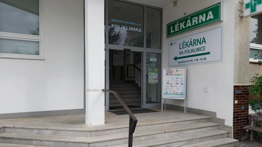 Vstup do budovy polikliniky s označením „Lékárna“. Schody vedoucí ke vchodu, informační tabule a cedule s provozní dobou lékárny umístěné vedle vstupních dveří.
